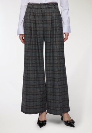 Donna che indossa pantaloni larghi a scacchi grigi con vita alta, abbinati a una camicia bianca a maniche lunghe e tacchi a punta neri con fiocchi.