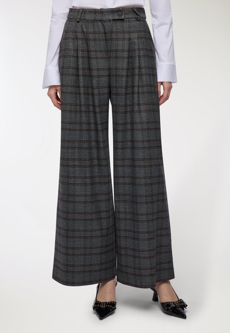 Donna che indossa pantaloni larghi a scacchi grigi con vita alta, abbinati a una camicia bianca a maniche lunghe e tacchi a punta neri con fiocchi.