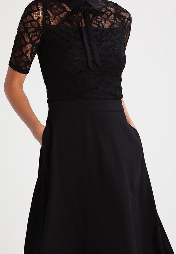 Femme portant un top en dentelle noire à manches courtes avec un détail noué au col, associé à une jupe noire taille haute avec poches.