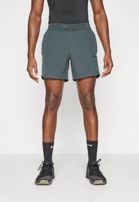 Zaļi sporta šorti ar melnu Nike logo un jostu, apvienoti ar melnām zeķēm un kedām, demonstrējot plūstošu, pieguļošu dizainu.