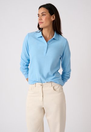 Femme en polo bleu clair à manches longues et pantalon beige, debout les mains dans le dos, regardant vers la gauche sur un fond uni.