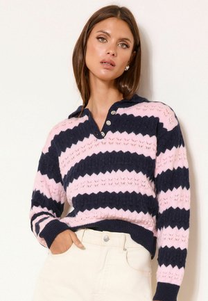 Frau trägt einen marineblauen und pink gestreiften Strickpullover mit Knöpfen am Kragen, kombiniert mit weißen, hoch taillierten Hosen.