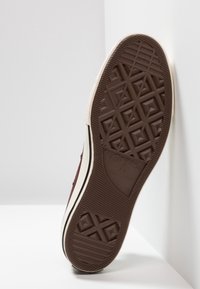 Suela de zapatilla de goma marrón con un patrón de relieve geométrico, una entresuela blanca y una franja lateral negra, diseñada para ofrecer agarre y durabilidad.