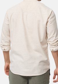 Mężczyzna ubrany w beżową koszulę z długim rękawem z podwiniętymi mankietami oraz zielone teksturowane spodnie, pokazany z tyłu na białym tle.