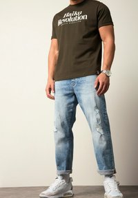 Camisa de manga corta verde oliva con texto blanco, jeans claros desgastados de color azul y zapatillas blancas con acentos grises y negros.