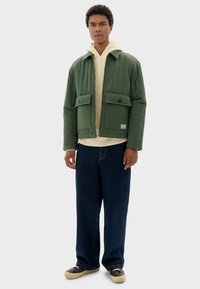 Gequiltede groene jas met twee voorkwamen, ritssluiting en hoge kraag, gedragen over een crèmekleurig sweatshirt en losse donkerblauwe spijkerbroek.