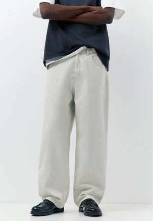 Persoană purtând pantaloni largi bej deschis, pantofi negri și o cămașă stratificată albă și bleumarin închis, cu brațele încrucișate pe un fundal simplu.