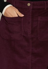 Jupe en velours côtelé bordeaux avec des côtes verticales, dotée d'une fermeture à pression, de deux poches avant et d'une surface texturée.