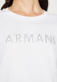 Valkoinen puuvillapaita, jossa on teksti "ARMANI EXCHANGE" kohotetuilla kirjaimilla, jotka on koristeltu hopeisilla niiteillä. Pyöreä kaula-aukko, lyhyet hihat, rento malli.