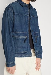 Jeansjacka i mörkblått, med knäppning framtill, två fickor med dragkedja och krage. Strukturen på tyget har en lätt urblekt finish.