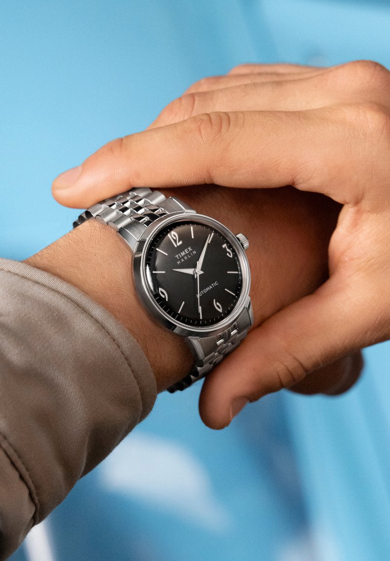 Person adjusting a silver Timex Marlin automatic wristwatch with a black dial and metal link band against a blue background.