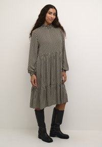 Robe longue noir et blanc en pied-de-poule avec un col haut, des manches volumineuses et une jupe à volants. Associée à des bottes noires montantes.