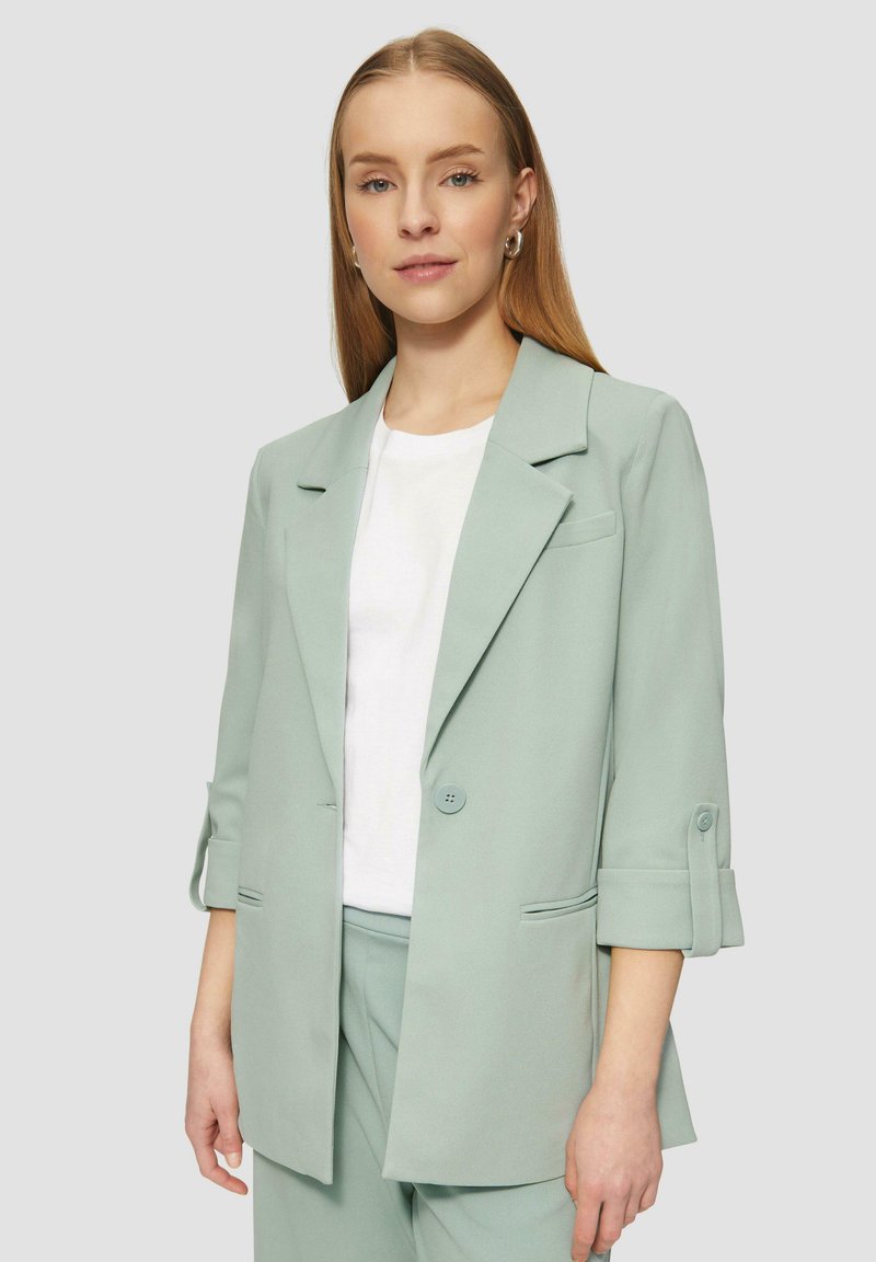 Hellgrünes, tailliertes Blazerjacke mit Reverskragen, einreihigem Knopfverschluss und hochgerolltem Ärmel, kombiniert mit einem weißen Oberteil darunter.