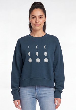Vrouw met donker haar in een paardenstaart, gekleed in een blauwe trui met illustraties van maanfases en lichtblauwe spijkerbroek, staand tegen een witte achtergrond.