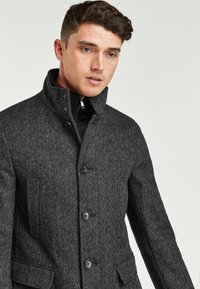Grahamuster Mantel in Grau mit hohem Kragen, ausgestattet mit vier vorderen Knöpfen, seitlichen Taschen und einer glatten Textur. Futter ist am Kragen sichtbar.