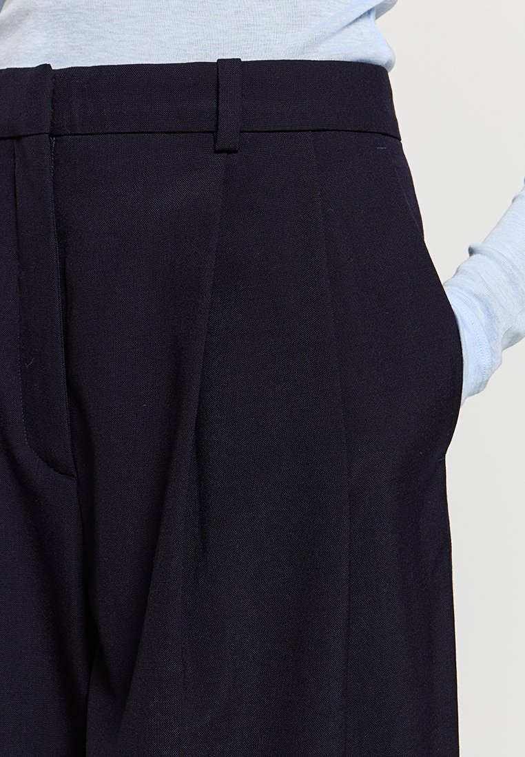 Granatowe, dopasowane spodnie z fakturowanej tkaniny, z przodu z fałdami i bocznymi kieszeniami, w połączeniu z jasnoniebieską koszulą z długim rękawem.