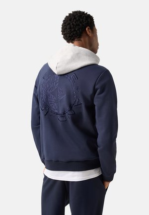 Homme portant un sweat à capuche bleu marine avec une capuche grise et un grand motif circulaire brodé dans le dos, debout devant un fond uni.
