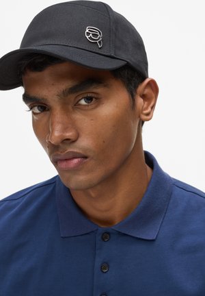 Young man wearing a navy blue polo shirt and black cap with a small metal pin shaped like sunglasses on the front.