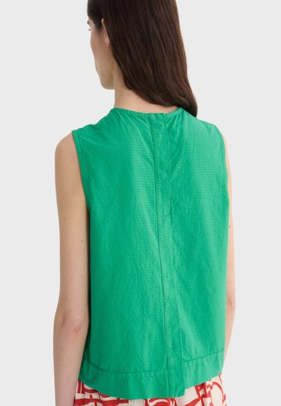 Femme aux longs cheveux bruns portant un haut vert sans manches et une jupe rouge à motifs, vue de dos sur un fond uni.