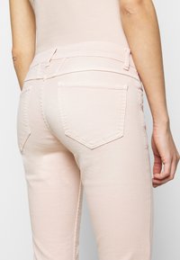 Jean pour femme rose clair avec une coupe ajustée, deux poches arrière et une texture de tissu lisse. Passants de ceinture visibles à la taille.