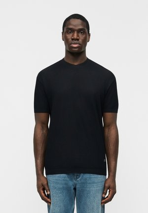 Homme portant un t-shirt noir à manches courtes côtelé et un jean bleu, debout devant un fond blanc uni, faisant face à l'avant avec une expression neutre.