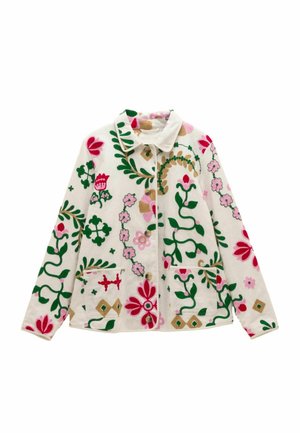 Hand teilweise in der Tasche eines weißen Stoffes mit grüner, pinker, roter und goldener Blumenstickerei am Jackenärmel und Körper.