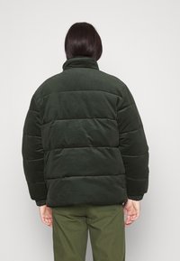 Chaqueta acolchada en verde oscuro, con secciones acolchadas, cuello alto y puños de canalé. Tejido suave y texturizado con un ajuste relajado.