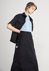 Black short-sleeved shirt layered over a light blue t-shirt, paired with a black skirt featuring cargo pockets and a logo patch. Accessorized with a striped bucket hat.