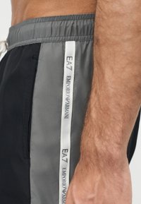 Gros plan sur le côté d'un homme portant un short noir et gris avec une bande verticale blanche portant l'inscription "EA7 Emporio Armani" près de la ceinture élastique.