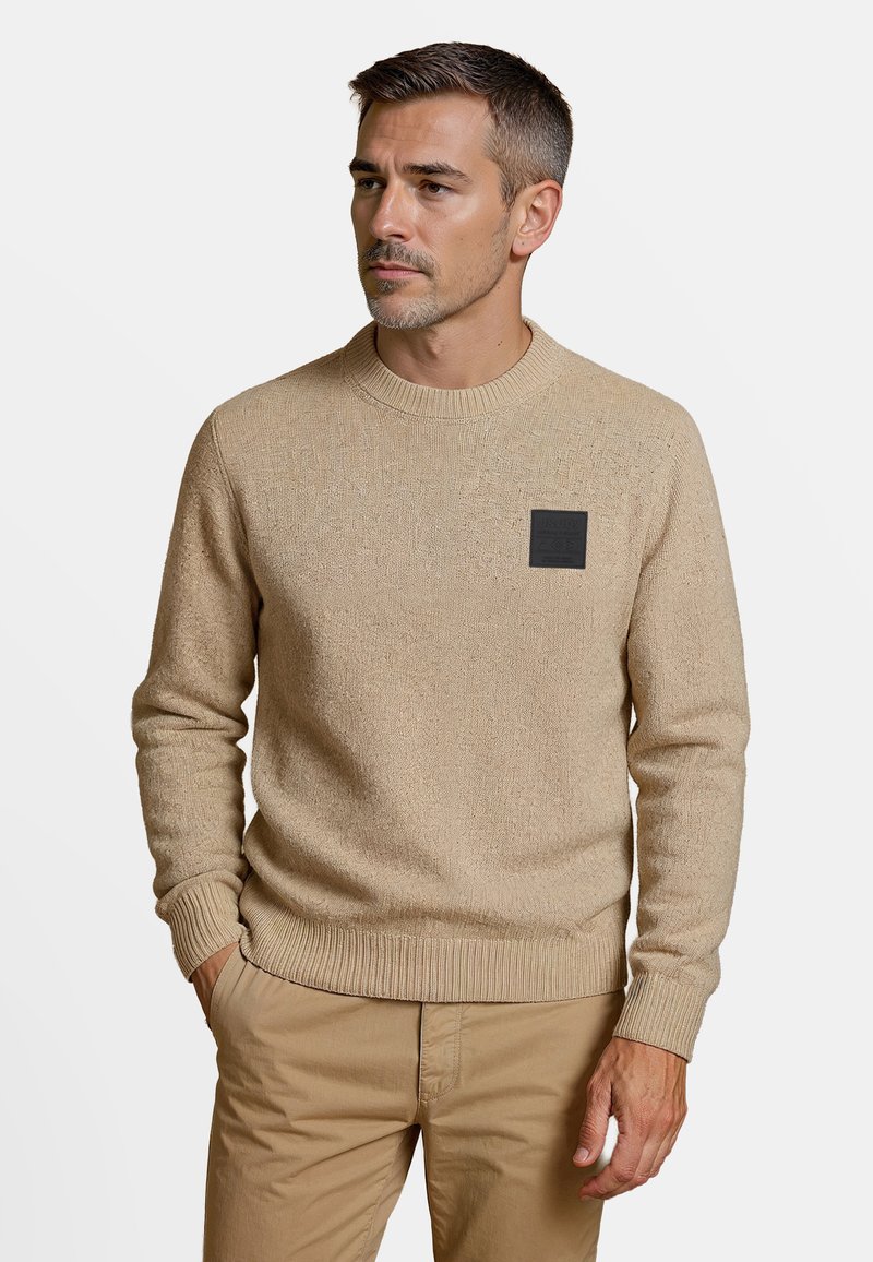 Beige gebreide trui met een ronde hals, textuurstof en een zwarte vierkante patch op de linkerkant van de borst; ribgebreide boorden en zoom.