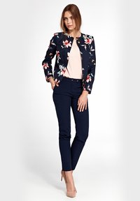 Kobieta stojąca z jedną ręką w kieszeni, ubrana w granatową kwiatową marynarkę, jasnoróżową bluzkę, wąskie granatowe spodnie i beżowe szpilki na białym tle.