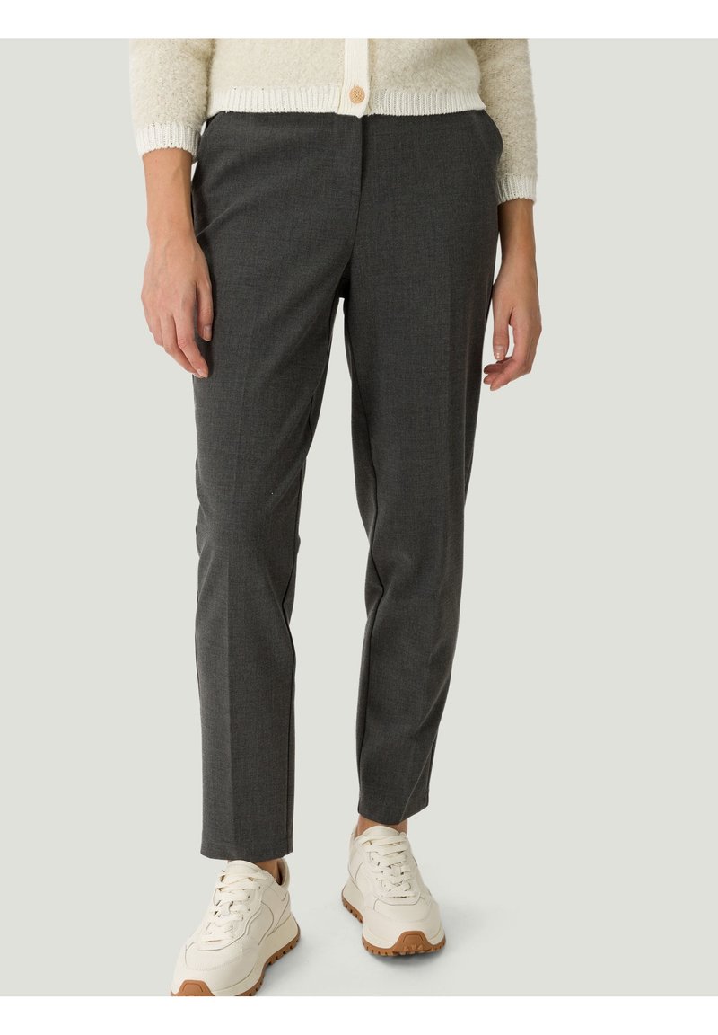 Femme portant un pantalon slim gris foncé, des baskets blanches et un cardigan crème avec un bouton en bois.