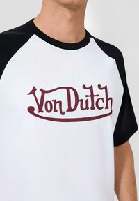 Bijela majica kratkih rukava s crnim raglan rukavima; sadrži bordo "Von Dutch" logo preko prsiju. Glatka pamučna tekstura.