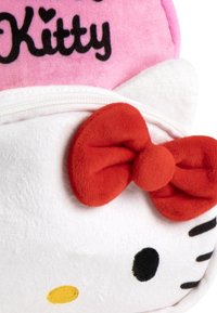 Visage en peluche blanc de Hello Kitty avec des yeux noirs, un nez jaune, un nœud rouge sur l'oreille, et un fond rose avec le texte "Kitty" en noir.