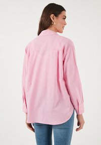Pink long-sleeve shirt with a back yoke, curved hemline, and buttoned cuffs. The fabric appears lightweight and slightly textured.