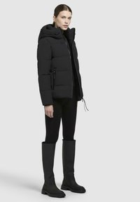 Veste rembourrée noire avec capuche, dotée d'une fermeture éclair à l'avant, de poches latérales et d'une silhouette ajustée. Associée à des leggings noirs et des bottes hautes.