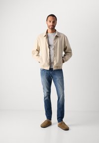 Jachetă bomber bej deschis, cu o textură netedă, tricou gri, blugi blue denim și pantofi slip-on maronii.