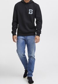 Sudadera negra con un bolsillo frontal y un logo azul claro en el pecho, combinada con jeans azul claro y zapatos negros con rayas blancas.