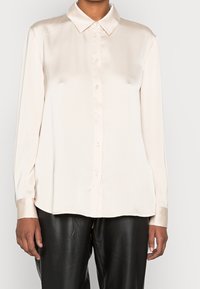 Blouse beige clair à manches longues en tissu satin, dotée d'un col classique et d'une patte de boutonnage sur le devant, avec un éclat subtil et une coupe ample.
