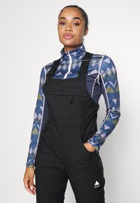 Sorte jumpsuit med justerbare stropper og forlomme, kombinert med en mønstrete langermet topp i flere farger som har geometriske design.