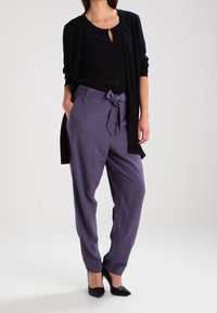 Svart cardigan över en svart topp, i kombination med högmidjade, lila byxor med detalj för knytning i midjan. Svarta högklackade skor fulländar outfiten.