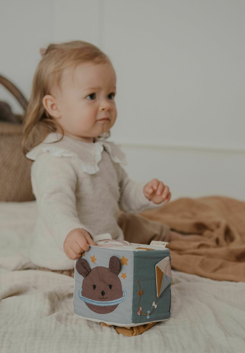 Un tout-petit aux cheveux châtain clair tient un cube en tissu doux orné de dessins d'animaux et d'étoiles, assis sur une couverture beige à l'intérieur.