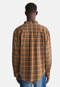 Brązowa i żółta kraciasta koszula z długim rękawem, z kołnierzykiem zapinanym na guziki i zaokrąglonym dołem. Posiada mankiety zapinane na guziki oraz szew yoke z tyłu.