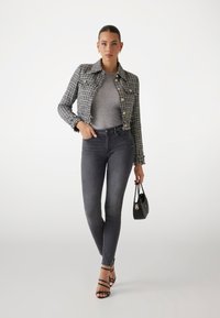 Zwart-wit tweed cropped jasje met gouden knopen, grijsgelakt top, grijze skinny jeans en zwarte sleehaksandalen, terwijl ze een klein zwart tasje vasthoudt.