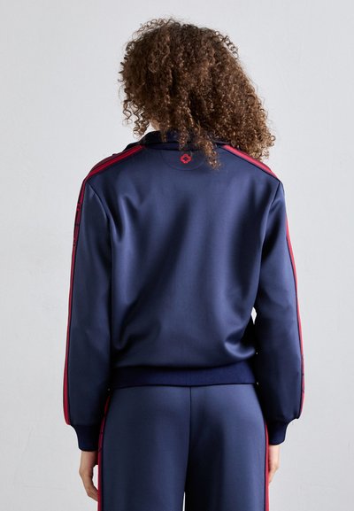 Giacca atletica blu navy con strisce rosse laterali, polsini e colletto a costine. Tessuto liscio con un dettaglio del logo nella parte posteriore del colletto.