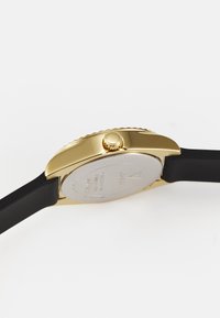 Reloj dorado con caja redonda, correa de silicona negra y corona texturizada. Cuenta con un fondo de acero inoxidable con detalles grabados.