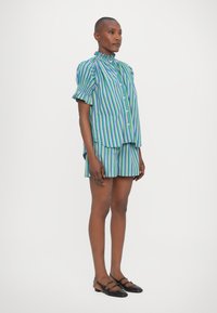 Conjunto de camisa de botones y pantalones cortos en rayas verticales azules, verdes y moradas; la camisa tiene un cuello con volantes y mangas cortas. Zapatos planos negros.