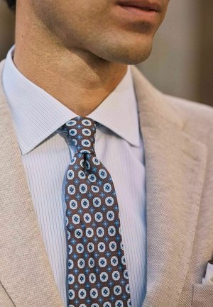 Mand iført beige tekstureret jakkesætjakke, lyseblå skjorte med nålestriber og brun slips med blå og hvide cirkelmønstre.