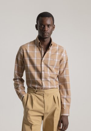 Camisa de cuadros marrón con cuello abotonado y detalles en blanco, combinada con pantalones beiges de cintura alta que presentan un botón frontal y pliegues.