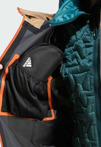 Unutrašnjost vodootporne jakne s tealcima nǎtvrtima, crna mrežasta džep u kojem se nalazi crni Adidas etui, narančasti detalji na džepovima.
